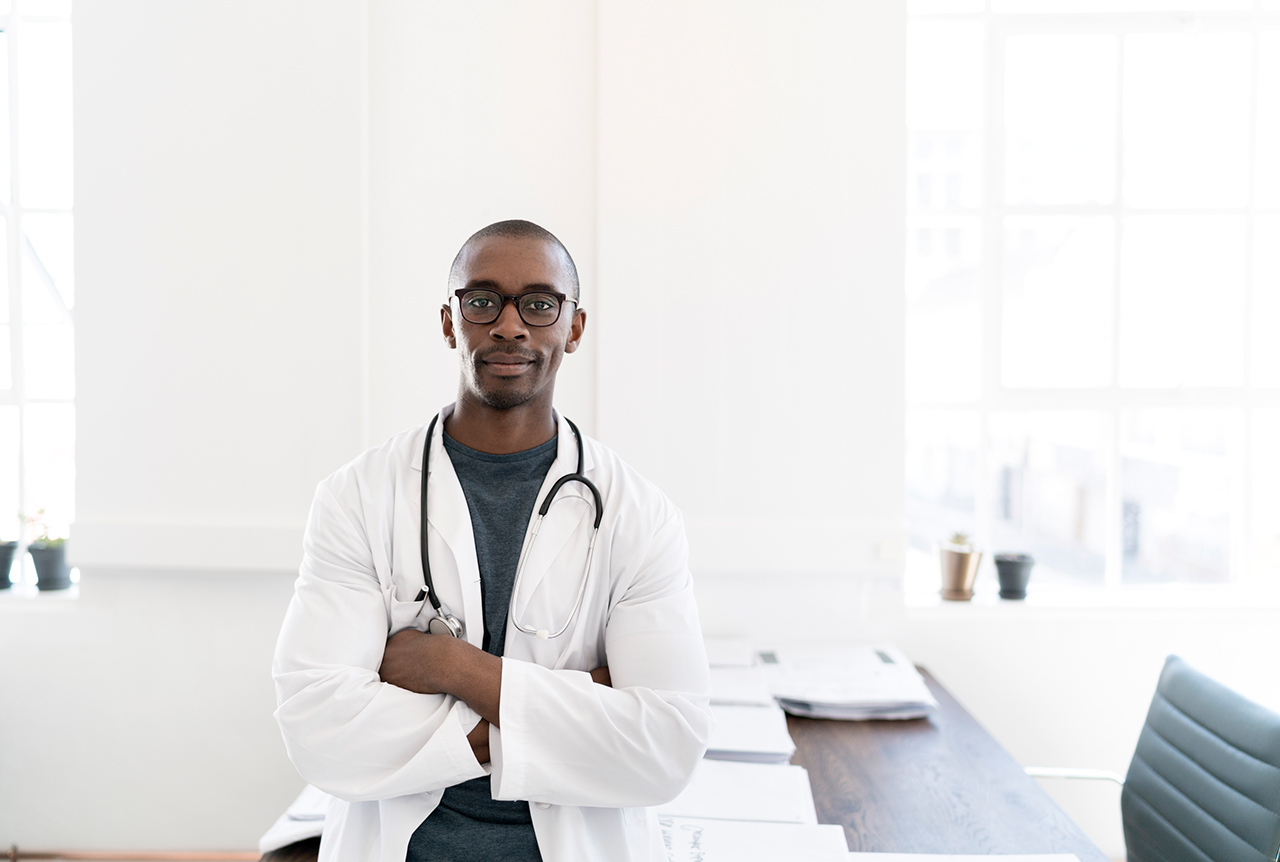 A photo of smiling male professional. Confident doctor wearing lab coat and stethoscope. He is standing with arms crossed.